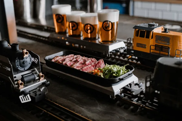 Teller mit Fleisch und Gemüse auf einer Miniatur-Eisenbahn, daneben Biergläser auf kleinen Waggons.