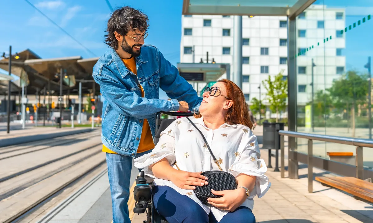 The image shows a person assisting another person in a wheelchair at a modern train station. In the background, there are tracks, a glass roof, and a multi-storey building.