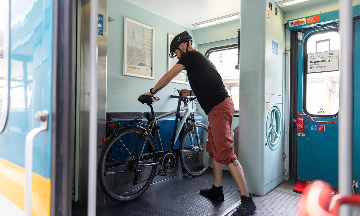 The image shows a person securing a bicycle in the multipurpose area of an alex train. On the door, there is a sign that reads “Regensburg – Munich.”