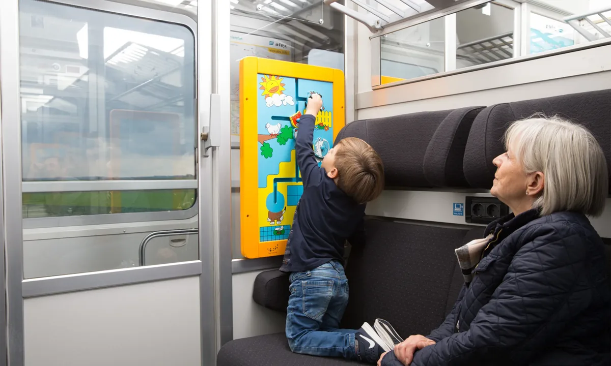 The image shows a child playing at a yellow activity wall inside an alex train. Next to the child, an adult is seated.