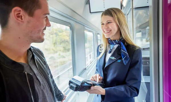 Obrázek zobrazuje osobu v uniformě, která používá mobilní zařízení k prodeji jízdenek ve vlaku alex. V pozadí jsou velká okna s výhledem na krajinu.