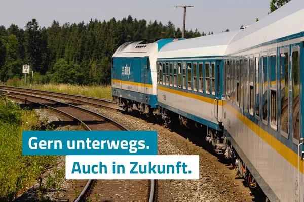 Ein blau-weiß-gelber alex-Zug fährt auf einer kurvenreichen Bahnstrecke durch eine grüne Waldlandschaft. Im Vordergrund verlaufen die Gleise, im Hintergrund sind Bäume und Vegetation zu sehen. Über dem Bild liegt der Schriftzug „Gern unterwegs. Auch in Zukunft.“, der die Fortführung des Bahnangebots symbolisiert.
