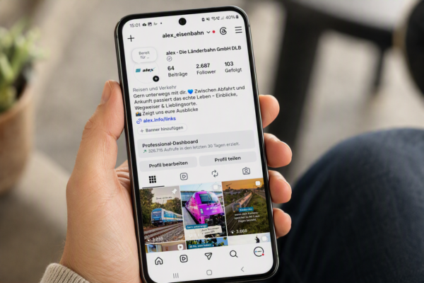 Eine Person hält ein Smartphone in der Hand. Auf dem Bildschirm ist das Instagram-Profil von alex zu sehen. Sichtbar sind das Profilbild, die Profilbeschreibung sowie mehrere Beitragsvorschauen mit Zügen und Reisethemen. Der Hintergrund des Bildes ist unscharf dargestellt und zeigt einen Innenraum.