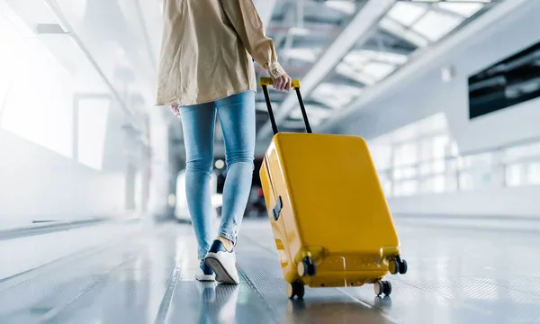 Obrázek zobrazuje osobu, která jde po světlé, moderní chodbě letiště a táhne za sebou velký žlutý kufr na kolečkách. Osoba je vidět zezadu, má světlou bundu, modré džíny a světlé boty. Okolí je tvořeno sklem a kovem, což podtrhuje prostředí letiště.