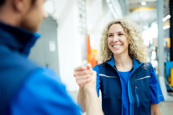 Zwei Personen in blauer Arbeitskleidung geben sich die Hand in der Werkstatt.