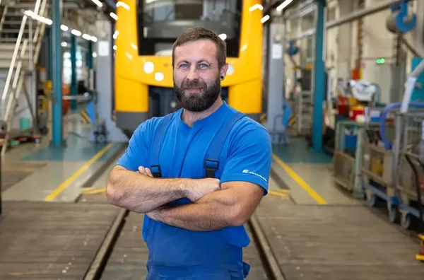 Person in blauer Arbeitskleidung steht mit verschränkten Armen vor einem gelben oberpfalzbahn-Zug.