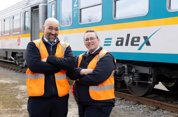 Zwei Personen in Warnwesten stehen vor einem alex-Zug neben dem Gleis.