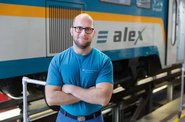 Person in blauer Arbeitskleidung steht mit verschränkten Armen vor einem alex-Zug.