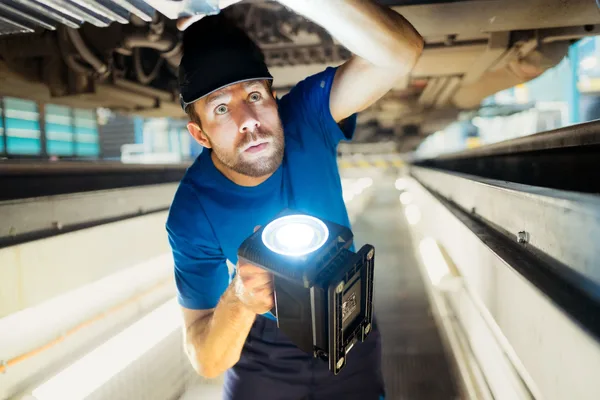 Person in blauer Arbeitskleidung inspiziert Bauteile unter dem Zug mit einer Lampe.