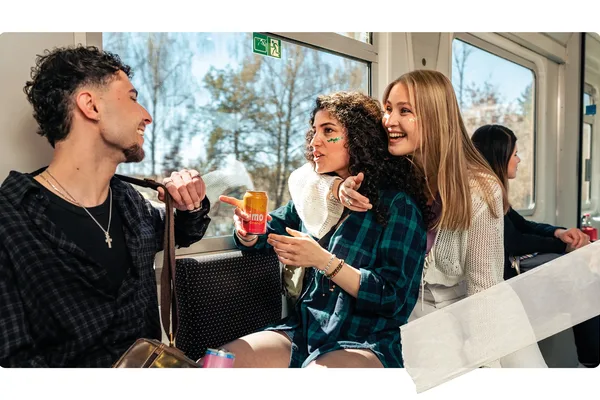 Eine Gruppe junger Menschen sitzt im Zug und unterhält sich. Ein Getränk wird gehalten, die Landschaft ist durch das Fenster sichtbar, während weitere Fahrgäste im Hintergrund sitzen.