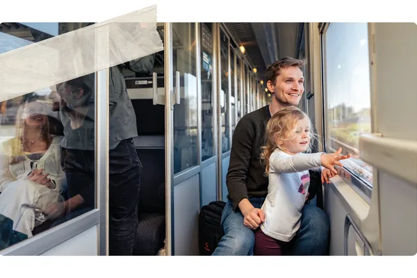 Ein Erwachsener sitzt mit einem Kind am Fenster im Zug. Das Kind zeigt nach draußen auf die vorbeiziehende Landschaft, während weitere Fahrgäste im Hintergrund zu sehen sind.