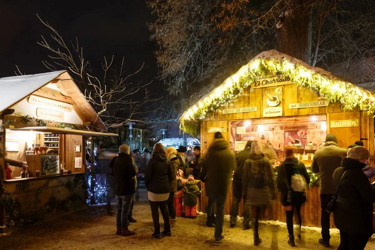 Stimmungsvoller Adventsmarkt mit beleuchteten Holzbuden, Menschen in Winterkleidung und festlicher Dekoration bei Nacht.