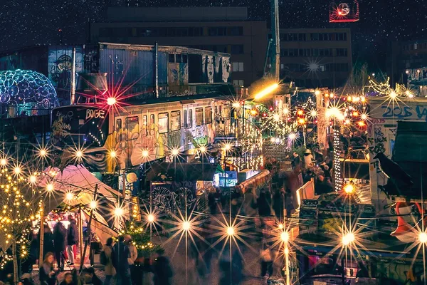 Bunt beleuchteter Zug inmitten eines alternativen Weihnachtsmarktes bei Nacht, umgeben von Lichterketten, festlicher Dekoration, Containern und Menschen in Bewegung.
