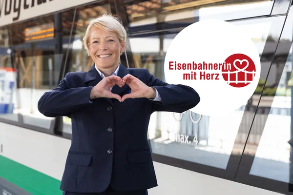 Rotes Herz-Schild mit der Aufschrift ‚Eisenbahner mit Herz‘ vor einem Bahnhof.