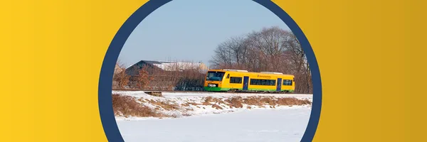 Ein gelb-grüner Triebwagen vom Typ Regio-Shuttle der oberpfalzbahn fährt an einem winterlichen Feldrand nahe Pösing entlang. Im Hintergrund stehen Bäume und einzelne Gebäude, der Boden ist schneebedeckt.