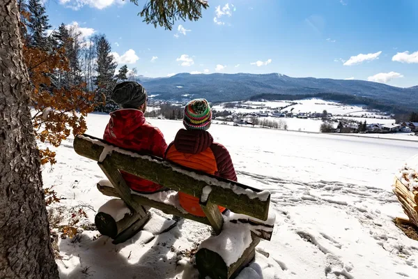 2 Wanderer im Winter auf einer Bank sitzend, die auf den schneebedeckten Ort Lam blicken.