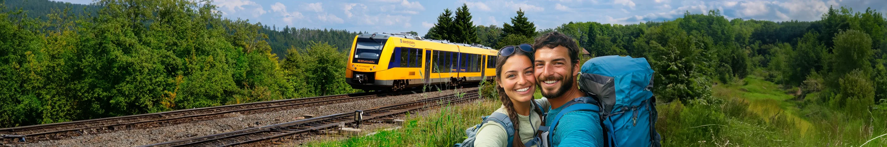 Zwei Personen vor einem gelben Zug der Länderbahn an einer Bahnstrecke im Grünen