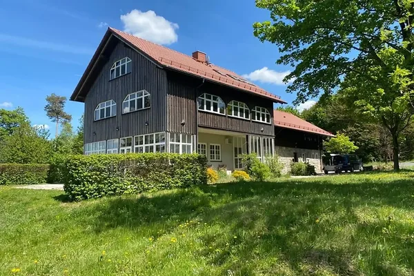 Das Bild zeigt das Marktredwitzer Haus, ein dreistöckiges Gebäude mit Holzfassade und Nebengebäude. Umgeben ist das Haus von grüner Wiese, Hecken und Bäumen.