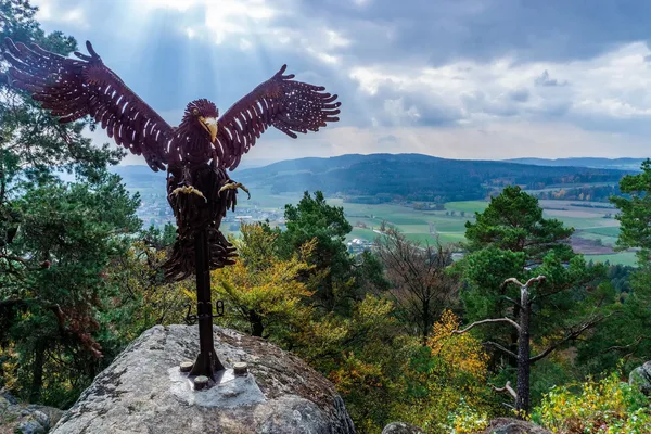 Das Bild zeigt eine Statue eines eisernen Adlers mit ausgebreiteten Flügeln auf dem Gipfel eines Bergs. Im Hintergrund ist ein Tal und weitere Berge zu sehen.
