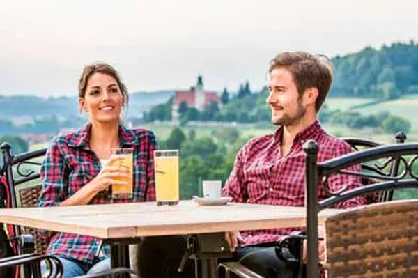 Zwei Personen genießen Getränke an einem Tisch im Freien, mit einer hügeligen Landschaft mit Ortschaft im Hintergrund