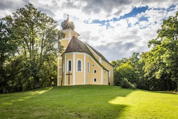 Das Bild zeigt die Kirche St. Barbara auf dem Eixlberg, die von grüner Wiese und Bäumen umgeben ist.