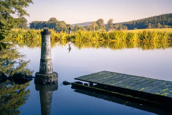 Ein Steg führt auf einen stillen See. Im Hintergrund ist Schilf und ein bewaldeter Hügel zu sehen.