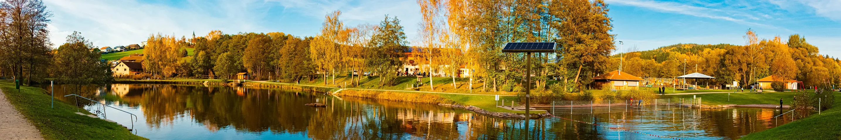 Blick auf einen See, an dessen anderem Ufer sich herbstliche Bäume und ein paar Häuser säumen. Ein Teil des Sees ist mit einer Absperrung abgetrennt.