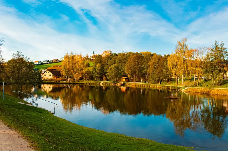 Blick auf einen See, an dessen anderem Ufer sich herbstliche Bäume und ein paar Häuser säumen. Ein Teil des Sees ist mit einer Absperrung abgetrennt.