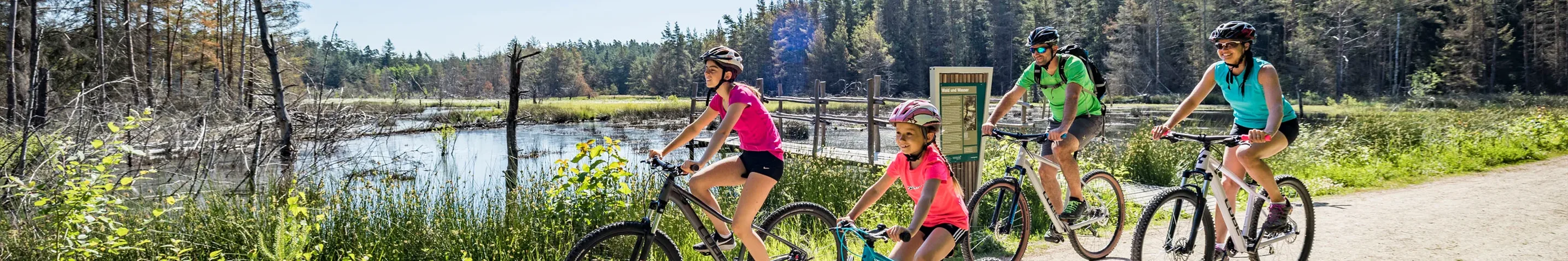 Eine Familie fährt mit ihren Rädern einen Kiesweg entlang. Im Hintergrund ist ein kleiner See zu sehen.