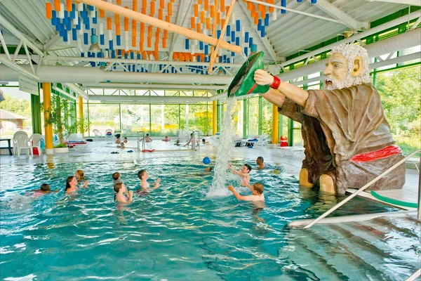 Kinder spielen im Wasser eines Hallenbads, daneben eine große Figur eines bärtigen Mannes, der einen grünen Eimer hält, aus dem Wasser fließt