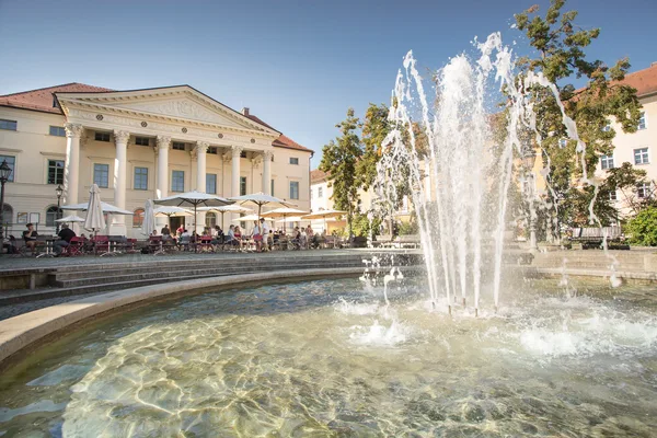Ein Brunnen mit Fontäne im Vordergrund. Im Hintergrund ist ein Gebäude mit Säulen und Tische, Stühle und Sonnenschirme auf einem großen Platz zu sehen.