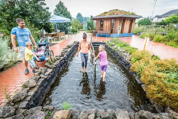 Mutter steht mit ihrer Tochter in einem flachen, natürlichen Wasserbecken, während der Vater mit Sohn und einem Kind im Kinderwagen vom Rand aus zusieht