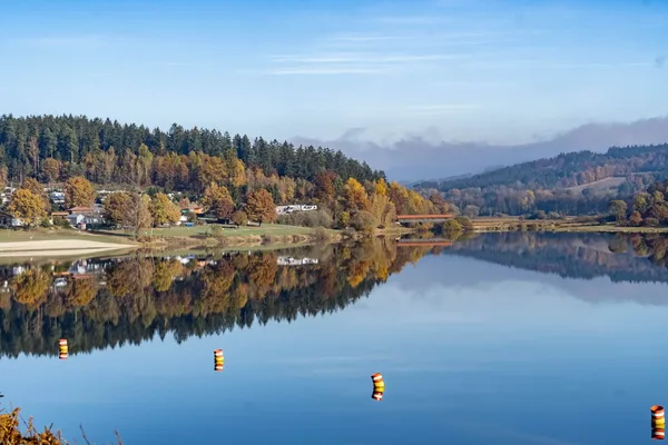 See in einer herbstlichen Landschaft mit bewaldeten Ufern und Hügeln im Hintergrund