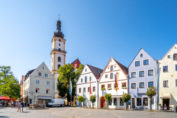 Ein Marktplatz mit hellen Häuserfassaden und einem Kirchturm im Sonnenschein