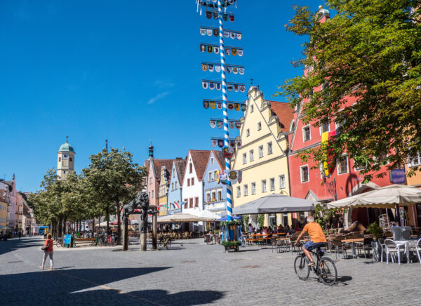Blick auf den Weidener Marktplatz, der von bunten Hausfassaden umgeben ist. Ein Fahrradfahrer fährt an den Freisitzen von Cafés vorbei und ein großer Maibaum mit vielen Wappen steht in der Mitte des Platzes.