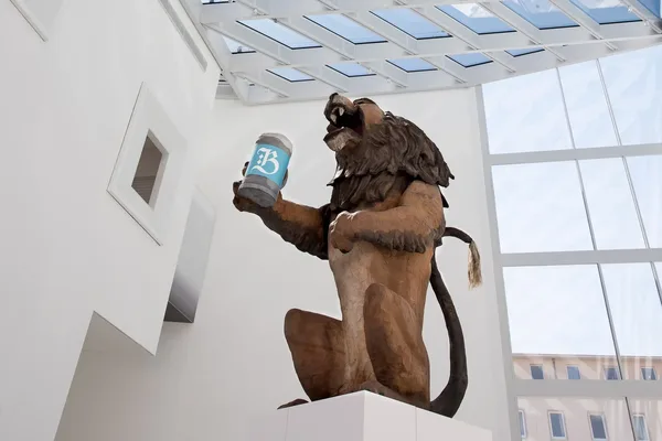 Eine Statue von einem Löwen, der einen steinernen Bierkrug hält, der mit einem großen weißen B auf hellblauem Hintergrund bedruckt ist.