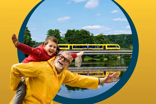 Ein älterer Mann in gelber Regenjacke trägt einen Jungen mit roter Regenjacke Huckepack. Im Hintergrund fährt die oberpfalzbahn an einem Fluss entlang, in dem sie sich spiegelt.