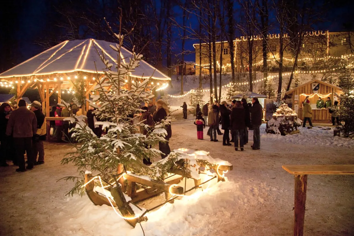 Ein Weihnachtsmarkt in der Dämmerung. Viele Buden und Wege sind beleuchtet. Es liegt Schnee.