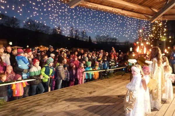 Auf einer Bühne stehen kleine Kinder in Engelskostümen. Im Publikum stehen weitere Kinder und Familien.