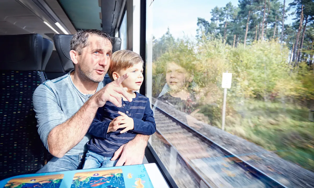 Erwachsene Person sitzt mit einem Kind am Fenster im Zug und betrachtet die vorbeiziehende Landschaft.