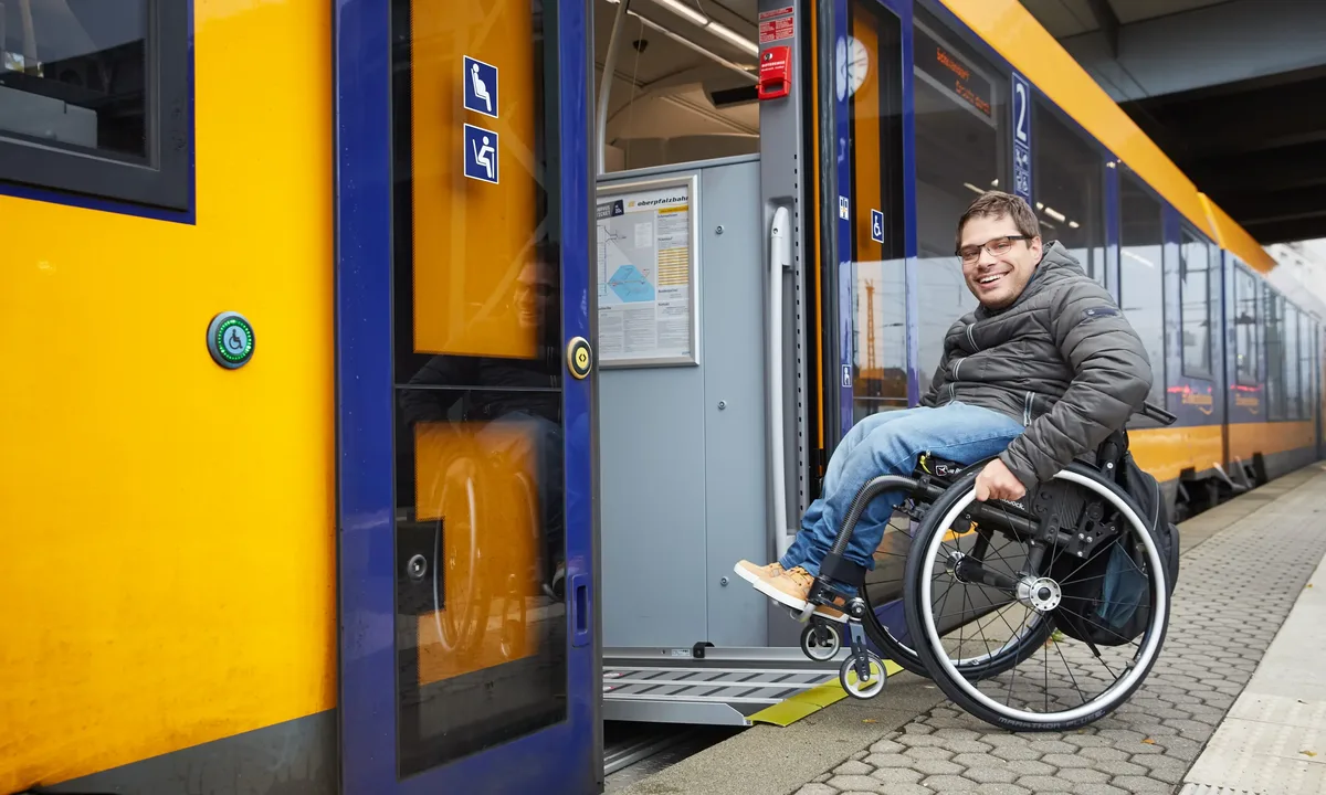 Person im Rollstuhl nutzt eine ausklappbare Rampe für den Einstieg in einen gelben oberpfalzbahn-Zug am Bahnsteig.