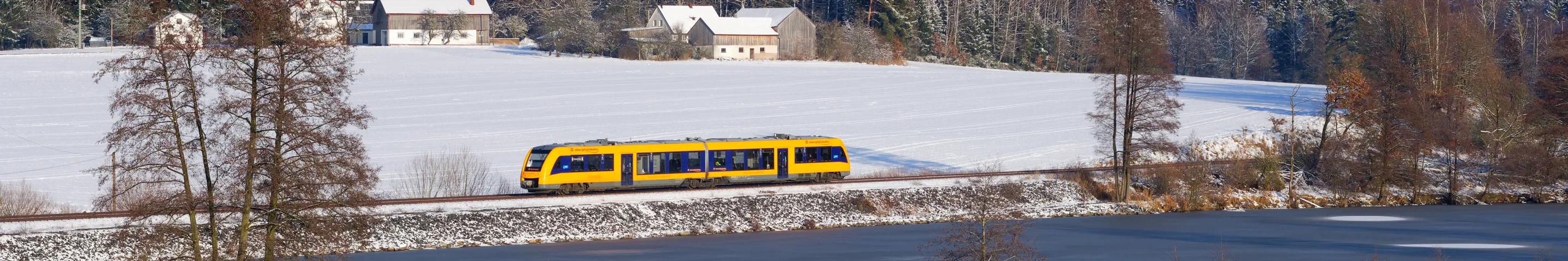 oberpfalzbahn-Zug auf schneebedeckter Strecke entlang eines zugefrorenen Gewässers, umgeben von Winterlandschaft.