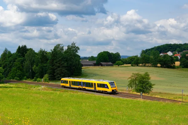 Gelb-blauer Lint-Triebwagen der oberpfalzbahn fährt durch das Naabtal in der Oberpfalz, umgeben von grünen Wiesen, Feldern und sanften Hügeln unter leicht bewölktem Himmel.