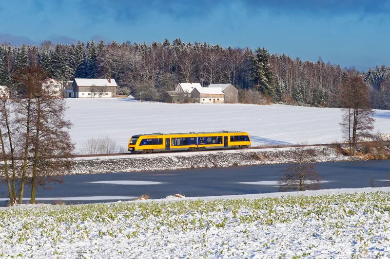 oberpfalzbahn-Zug auf schneebedeckter Strecke entlang eines zugefrorenen Gewässers, umgeben von Winterlandschaft.