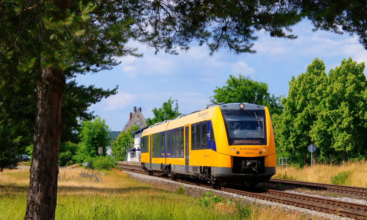 Gelber oberpfalzbahn-Zug fährt durch grüne Landschaft mit Bäumen und einem Gebäude im Hintergrund bei sonnigem Wetter.