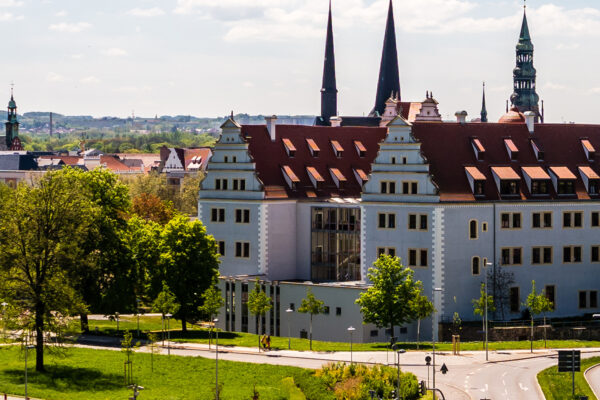 Historisches Gebäude in Zwickau mit heller Fassade und roten Ziegeldächern, umgeben von Grünflächen und Bäumen: August Horch Museum. Im Hintergrund prägen mehrere Kirchtürme die Stadtsilhouette, während im Vordergrund Straßen und Wege durch eine parkähnliche Umgebung führen.