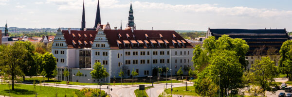 Historisches Gebäude in Zwickau mit heller Fassade und roten Ziegeldächern, umgeben von Grünflächen und Bäumen: August-Horch Museum. Im Hintergrund prägen mehrere Kirchtürme die Stadtsilhouette, während im Vordergrund Straßen und Wege durch eine parkähnliche Umgebung führen.