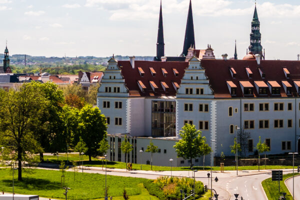 Historisches Gebäude in Zwickau mit heller Fassade und roten Ziegeldächern, umgeben von Grünflächen und Bäumen: Das August-Horch-Museum. Im Hintergrund prägen mehrere Kirchtürme die Stadtsilhouette, während im Vordergrund Straßen und Wege durch eine parkähnliche Umgebung führen.