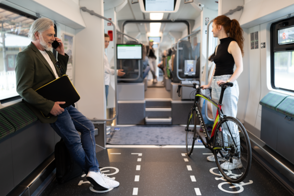 Zu sehen ist der Mehrzweckbereich im Siemens Mireo. Links ein mittelalter Mann mit Laptoptasche und telefoniert, recht m Bild steht eine junge Frau mit Fahrrad. Im Hintergrund sind weitere Fahrgäste zu sehen.