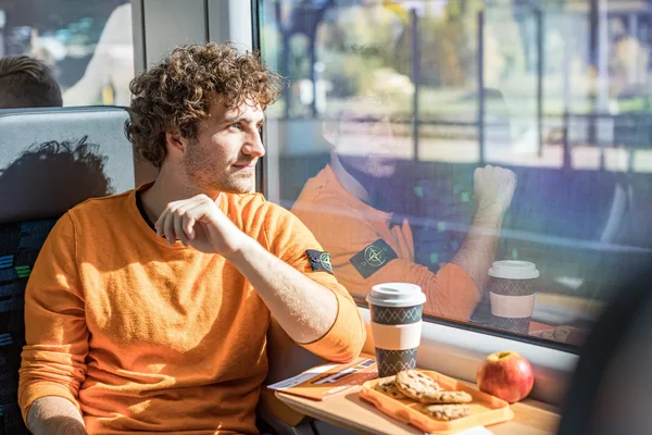 Ein Reisender sitzt am Fenster mit Blick aus dem Zug hinaus. Er trägt einen orangen Pullover und vor ihm steht ein oranger Kaffeebecher.